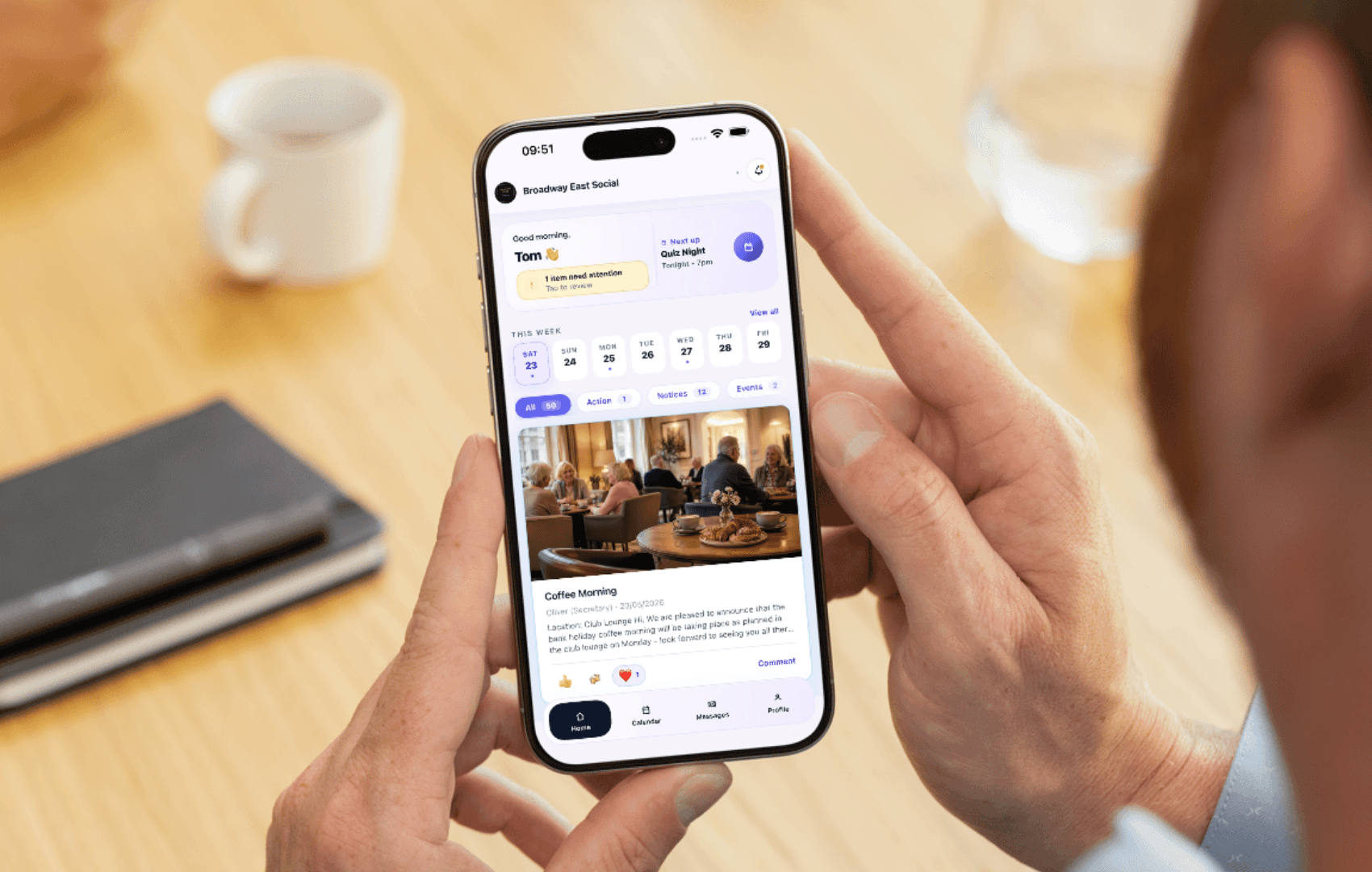 A club member holding an iPhone at a table with a coffee and notebook, viewing the Comnly member feed showing a morning greeting, a Quiz Night next-up card and a Coffee Morning post.